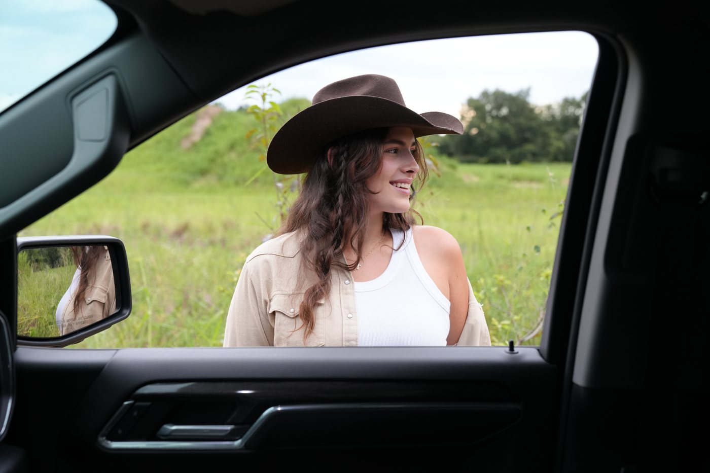 Flora in a cowboy hat, smiling, seen through the open window of a truck in a green Florida field.
