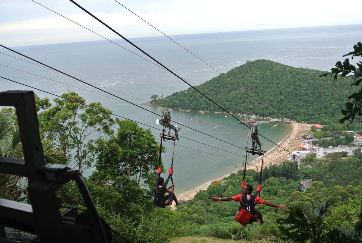 Two people ziplining over a green bay and beach in Brazil.