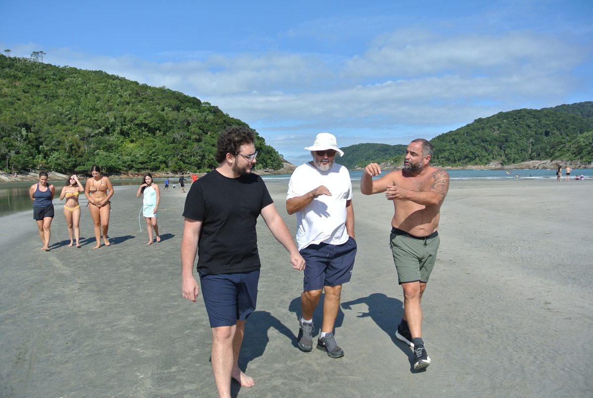 Three men laughing as they walk along a beach between green hills.