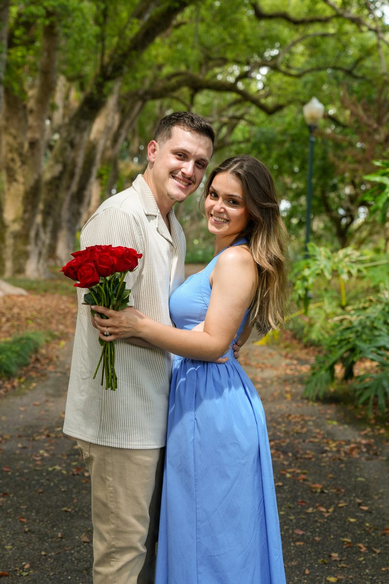 The couple smiling on an oak-lined path, holding red roses.
